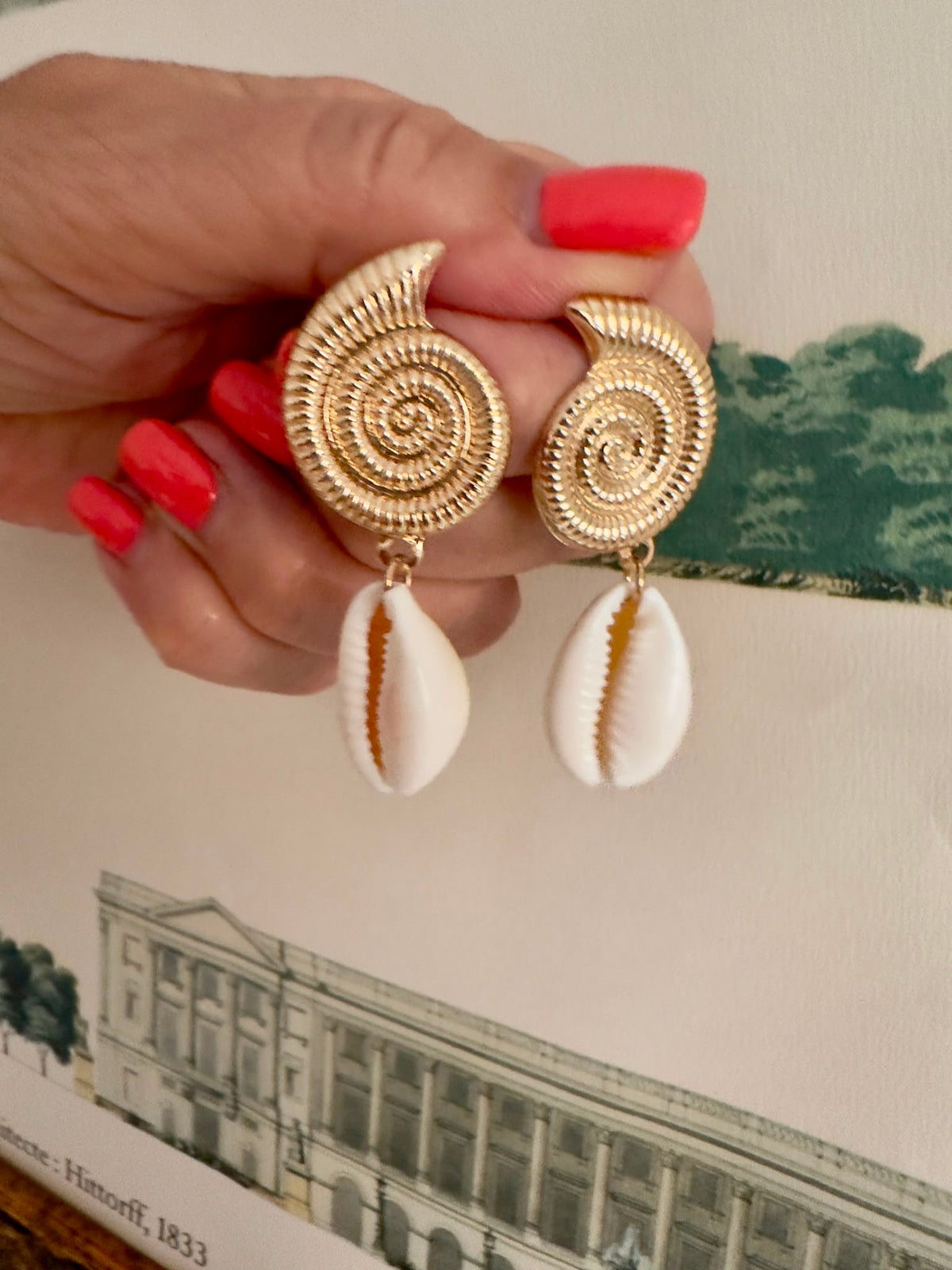 Pendientes Caracola Dorada en mano, detalle de la espiral dorada y concha cowrie natural